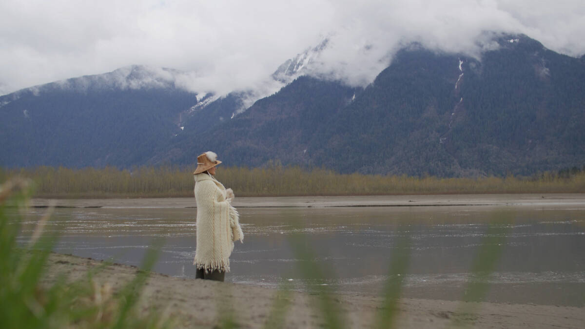 A person wearing a traditional Indigenous hat and shawl stands on the banks of the Fraser River with snow dusted mountains in the background.