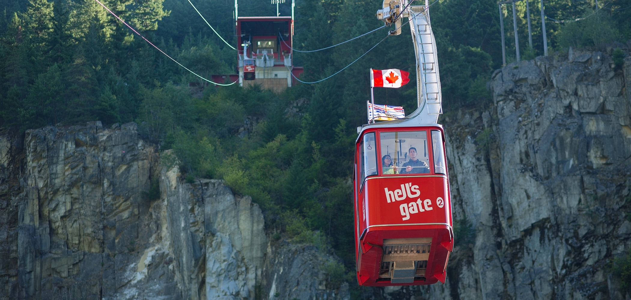 A red tram flying a BC and a Canada flag crosses a gorge with a rocky cliff at the top of which sits a structure where the trams load and offload.