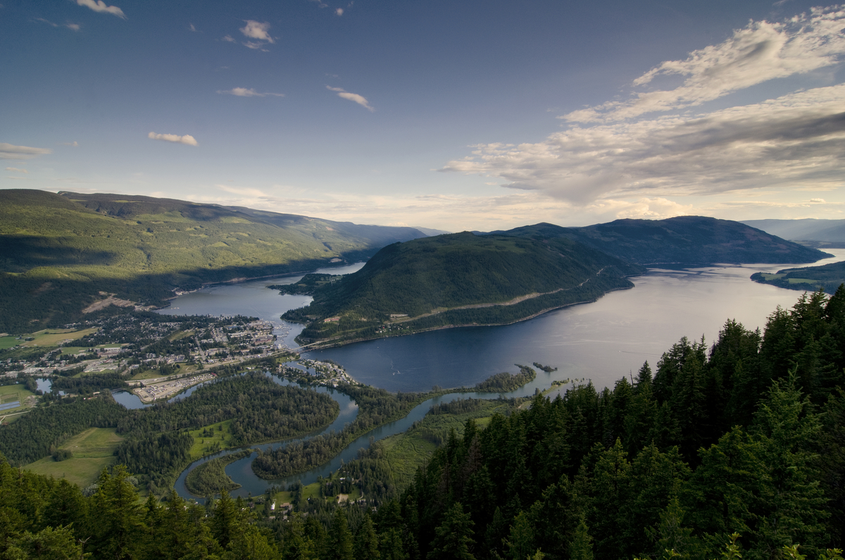 An aerial shot of Shuswap Lake during the day time. A small town is seen on one side surrounded by rolling green hills and the blue lake.