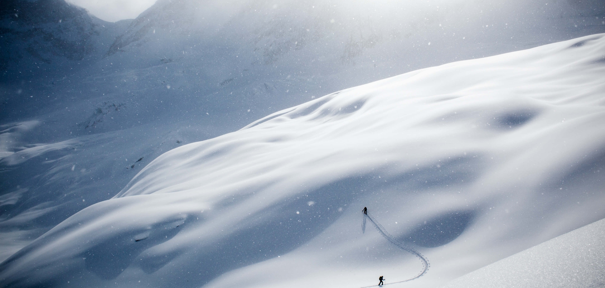 A person hikes up a steep snowy ski slope in the alpine. It is snowing heavily.