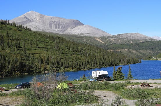 Campers and tents are set up at the edge of the water, overlooking a dense forest and a rocky mountain.