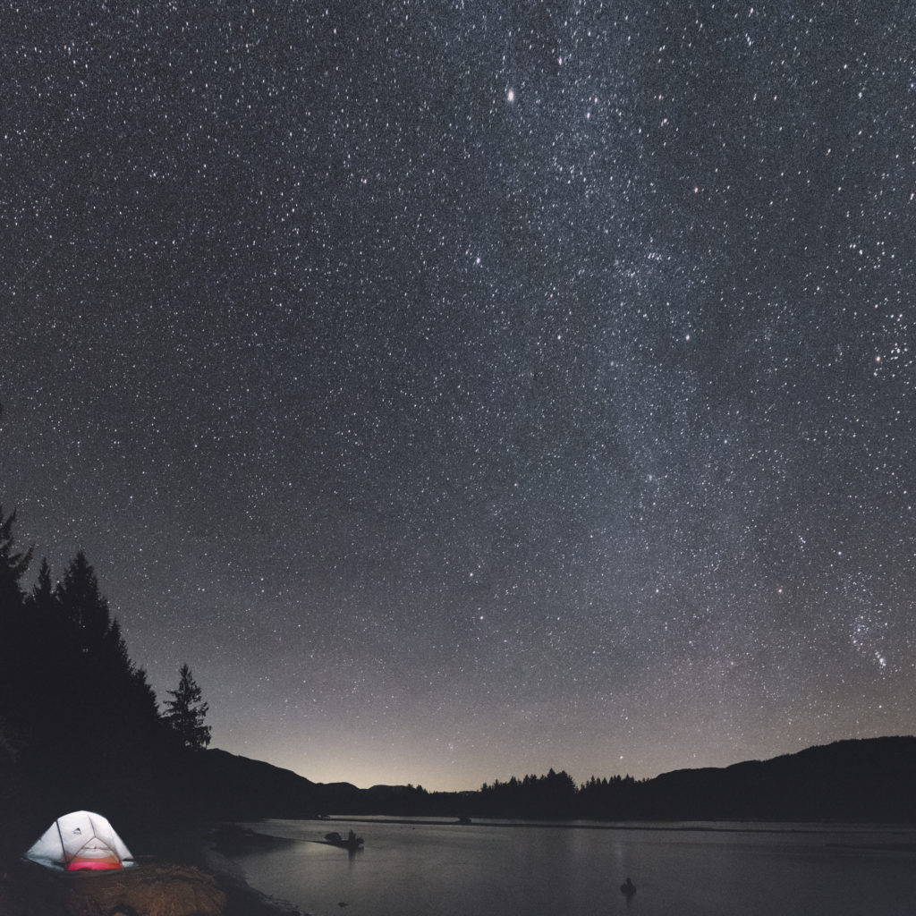 A brightly lit tent is pitched at the edge of the water under a starry sky.