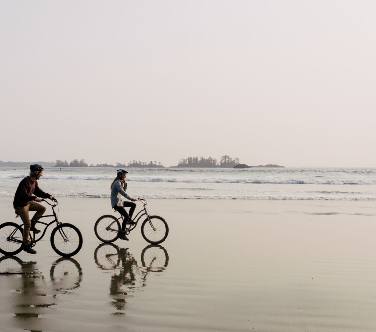 Couple riding bikes along the sand with the ocean and small islands off the coast behind them
