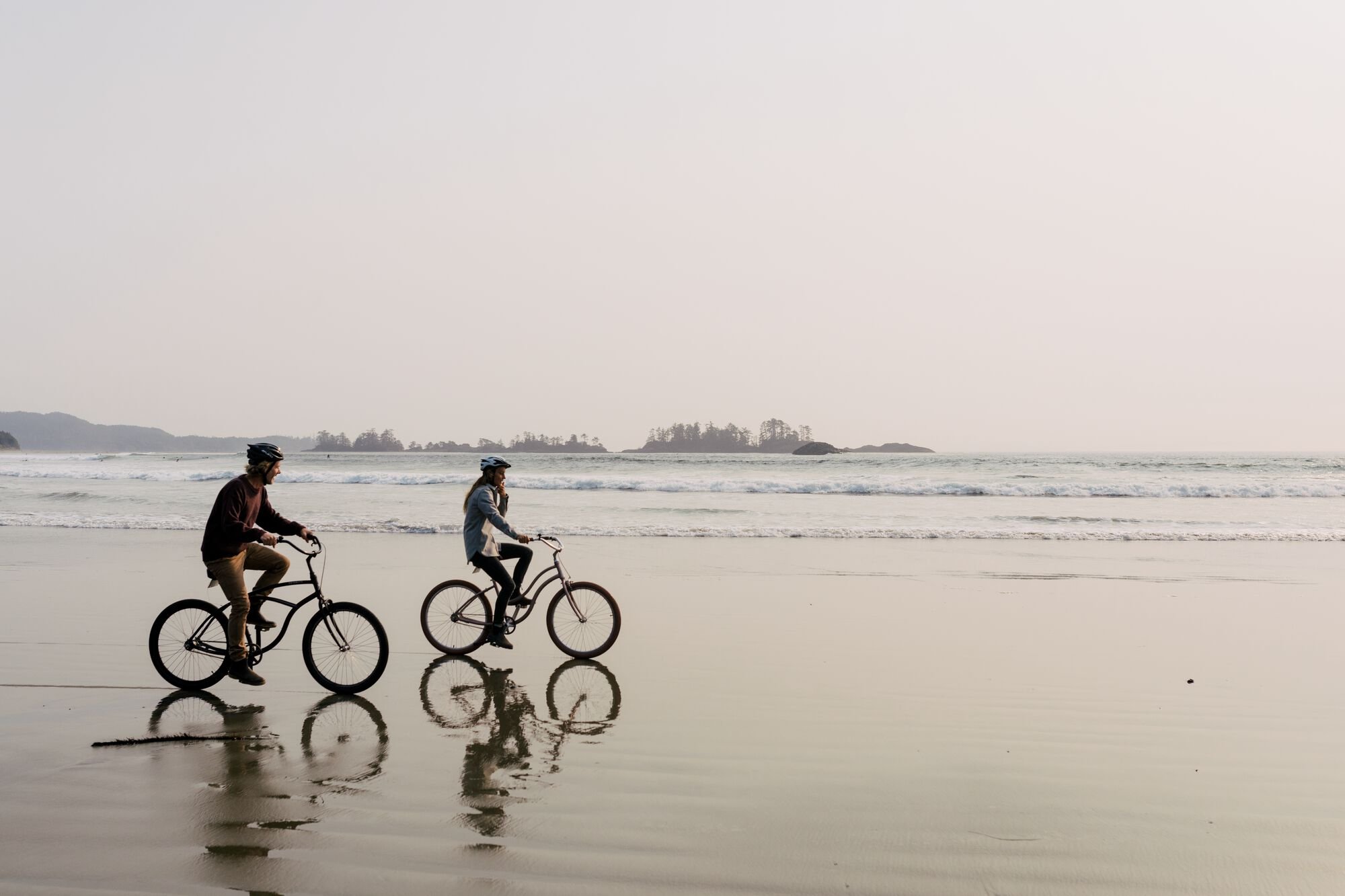 Couple riding bikes along the sand with the ocean and small islands off the coast behind them