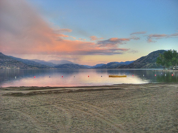 Skaha Lake with the sandy beach in the foreground and glass-like lake in the background and a pink sunset above.
