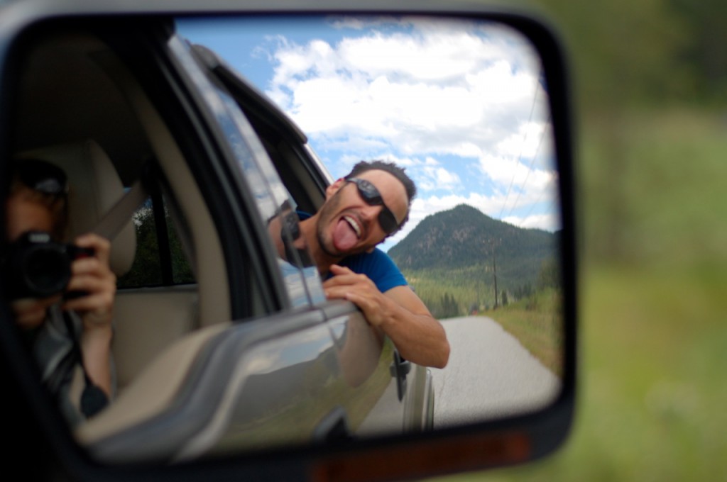 A man sticks his head out of a car window and sticks his tongue out in the reflection of the side mirror.