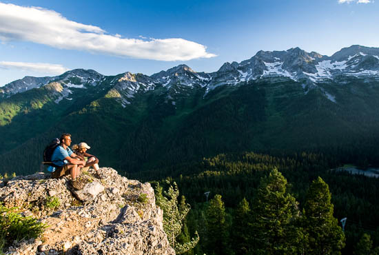 Two hikers sit at the edge of a cliff, taking in the views of the snow-capped mountains.