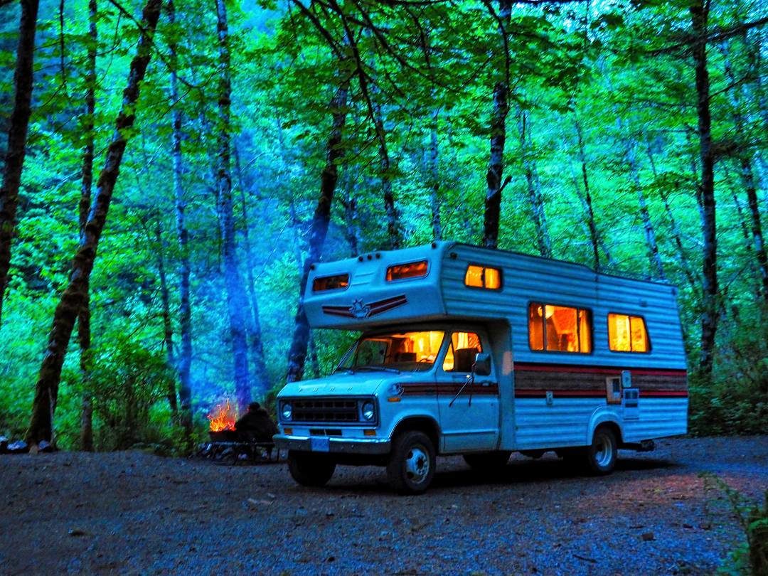 An RV is parked in a campsite, next to a roaring campfire at dusk.