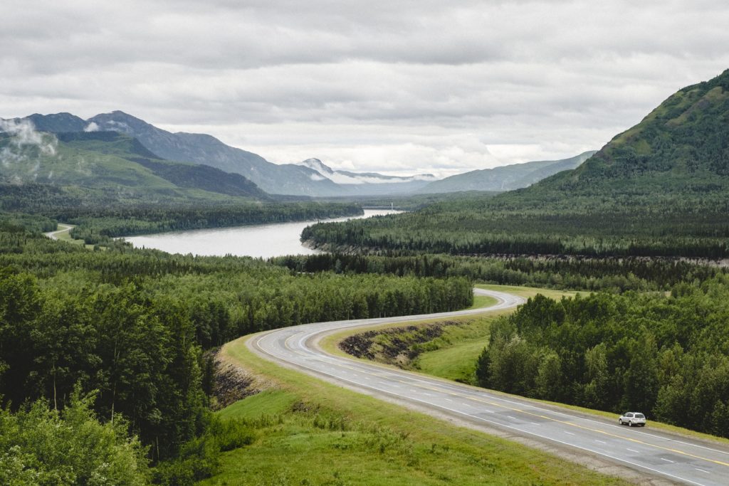 A highway winds through the dense wilderness, and past a lake nestled at the bottom of a mountain range.