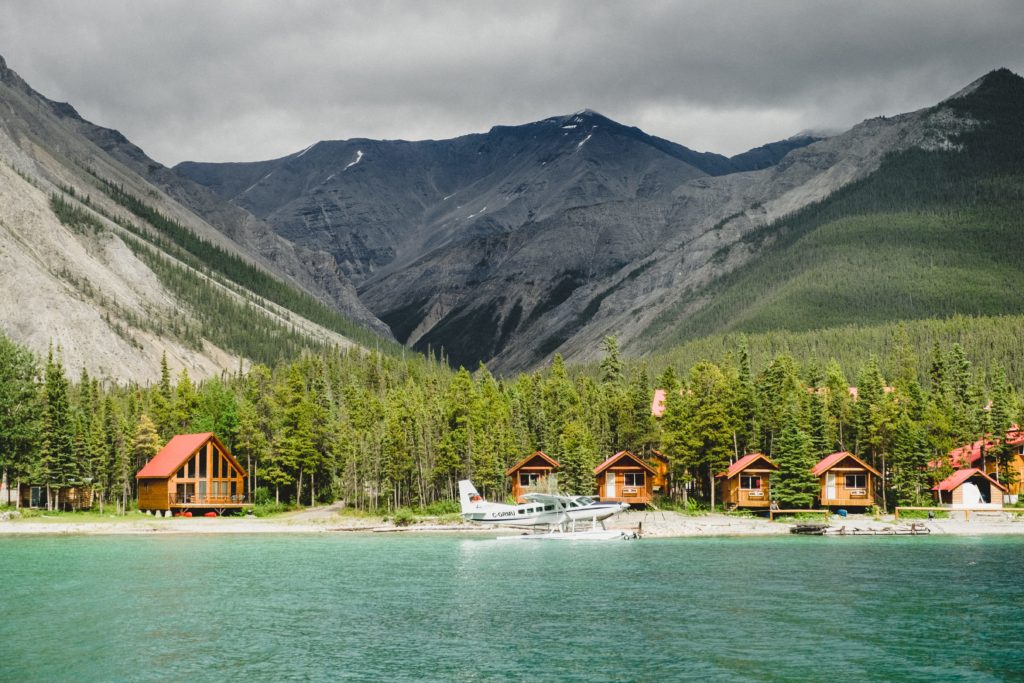 A seaplane docked at a beach lined with wood lodges nestled at the base of a rocky mountain range.