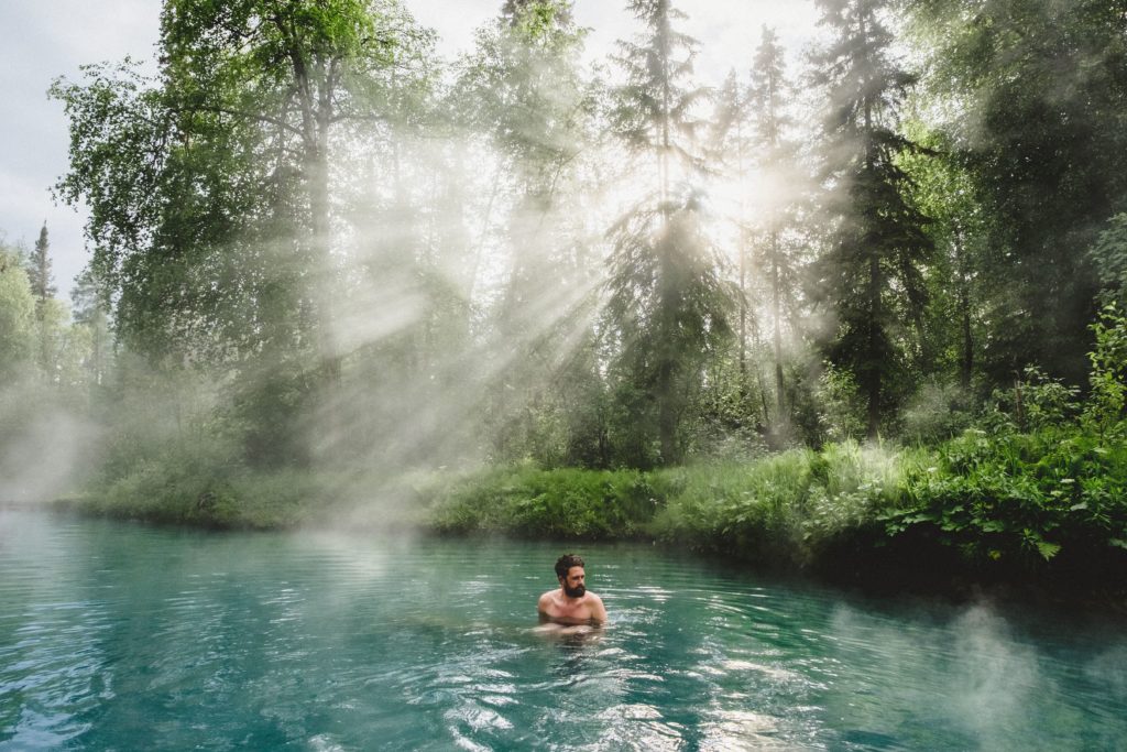 A man relaxes in a turquoise hot spring drenched in sunlight.