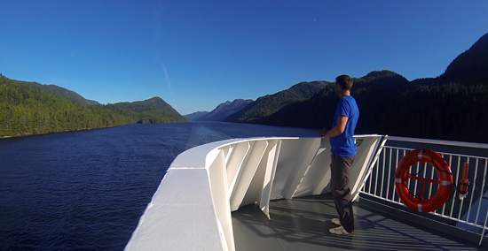 A man takes in a mountain landscape from the deck of a ferry.