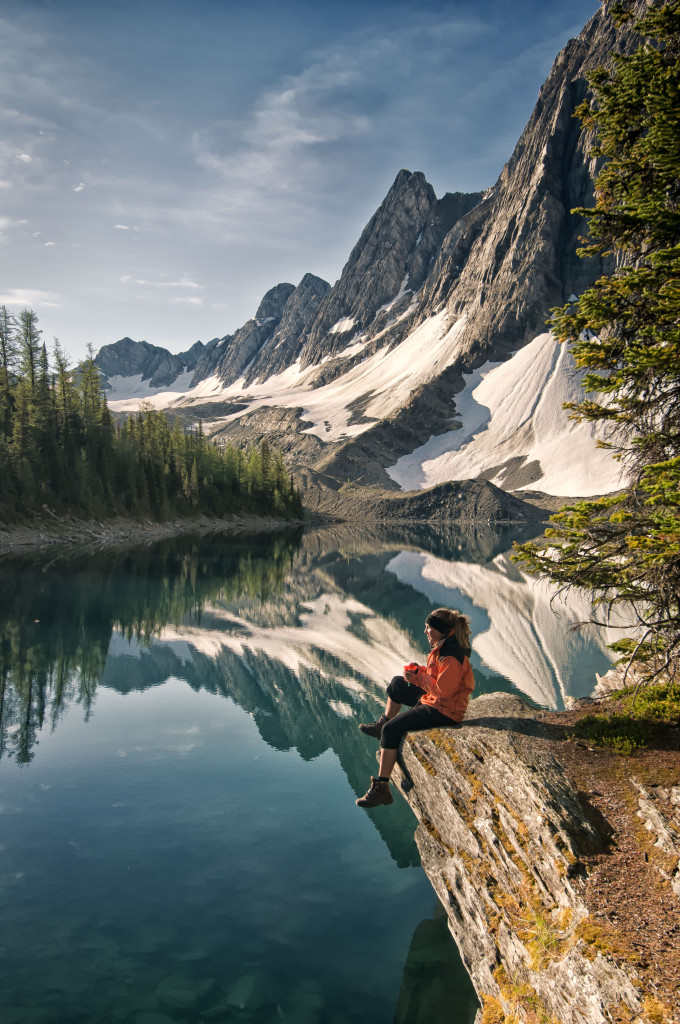 A hiker sits on the edge of a cliff, over placid waters and next to snow-capped mountains.
