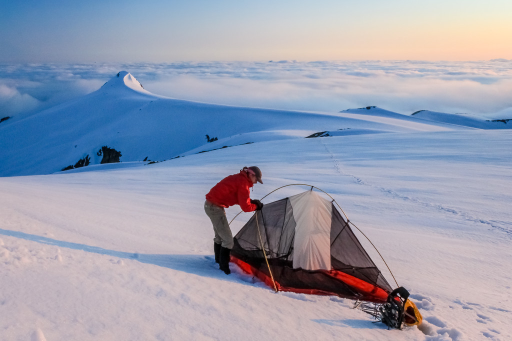 A woman pitches a tent at the summit of a snow covered mountain.