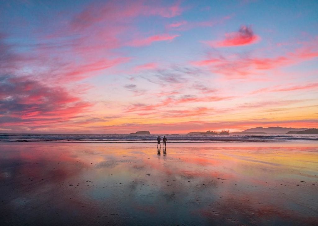 A couple stroll a quiet beach under a pink and orange sunset.