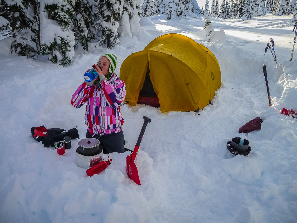 A woman sits in the snow outside her tent, drinking from a water bottle next to a red shovel and a small camping stove. 