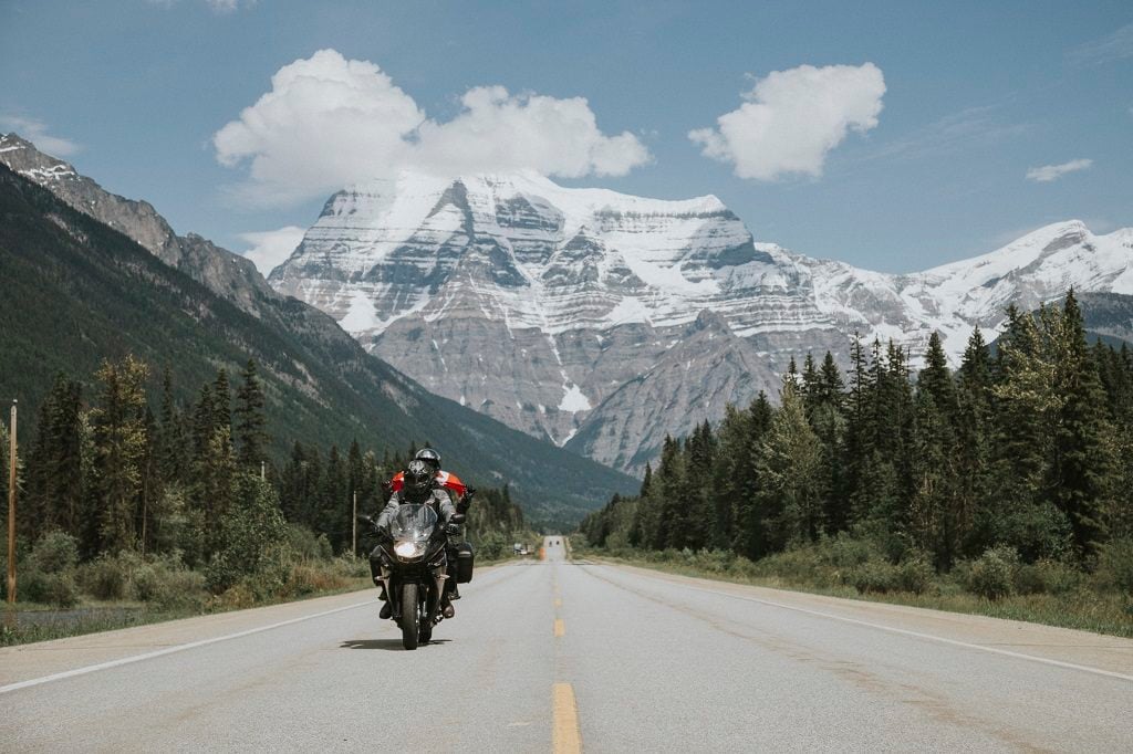 A motorcycle travels down the highway, with snow-capped mountains in the background.