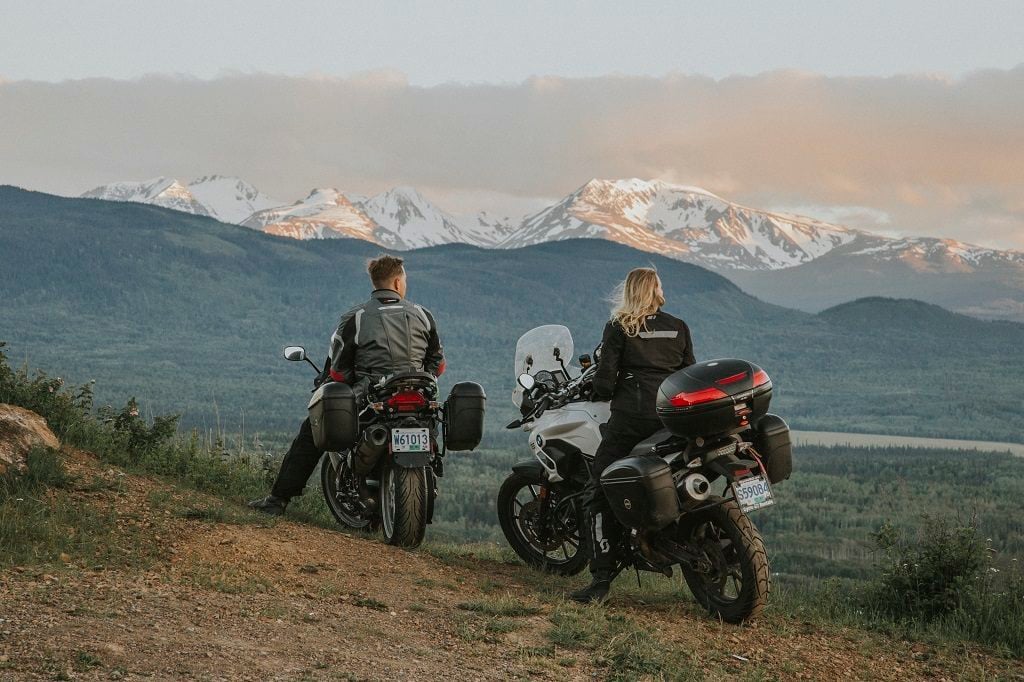 A woman and a man pull over on their motorcycles to take in the view of snow-covered mountains.