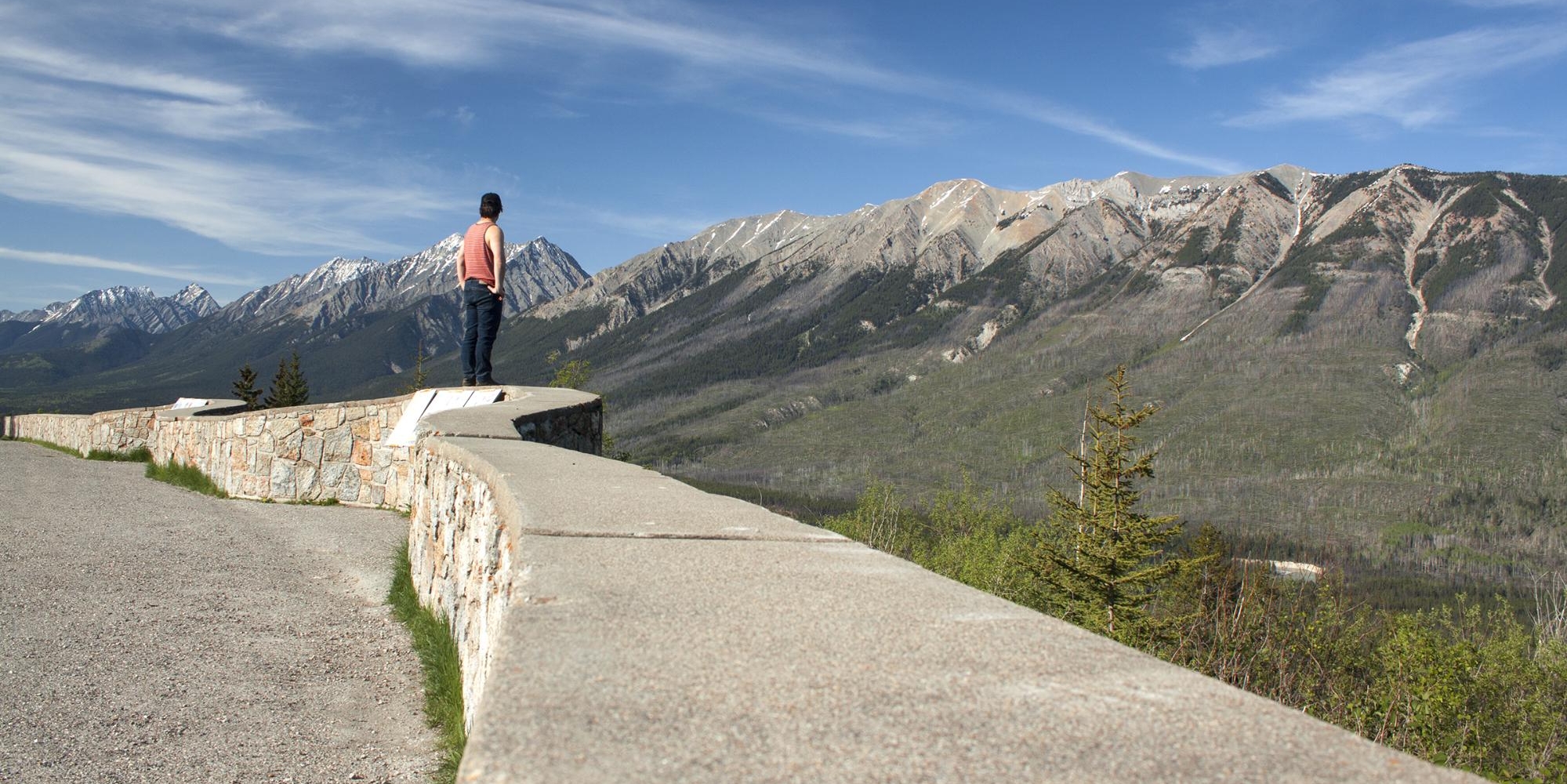 A man stands at the edge of a lookout, taking in a rocky mountain range.