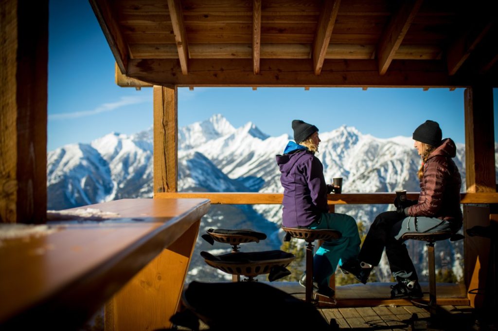 Two woman in ski jackets enjoy coffee on an outdoor patio with views of snow-covered mountains.