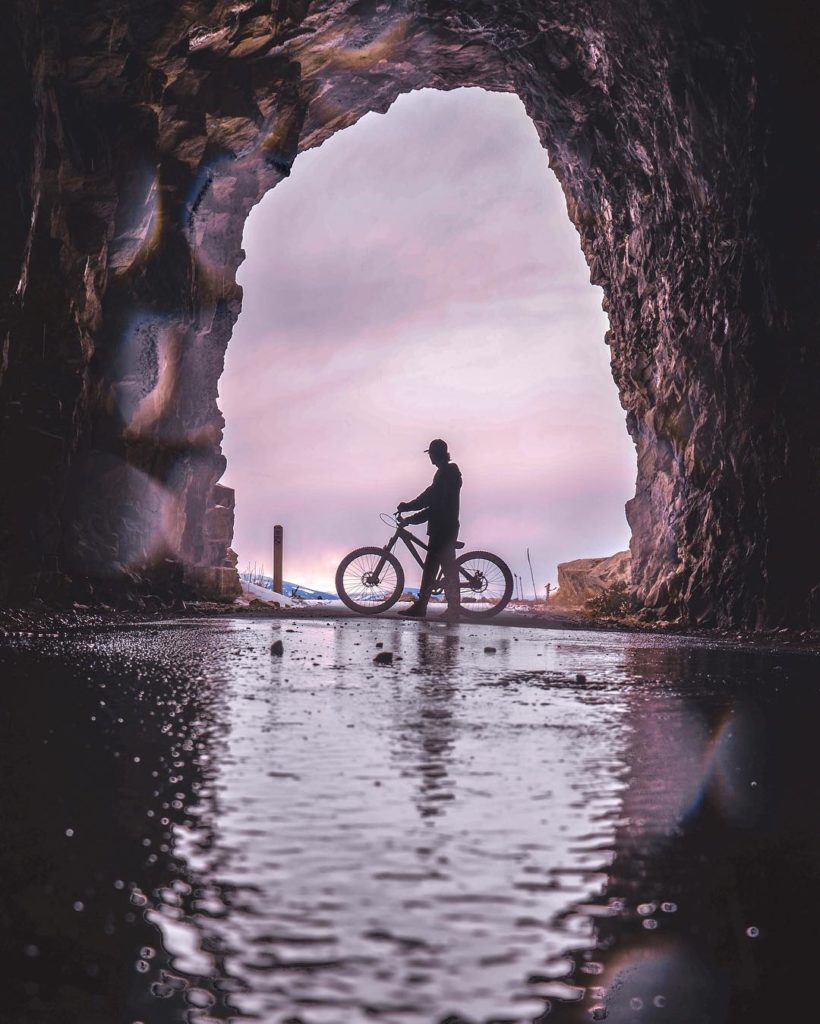 A man walks his bicycle past tunnel at sunset.
