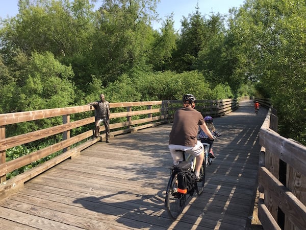 Three bikers (one man and two children) biking across the wooden trestle bridge on the Lochside Regional Trail, with a statue of the Saanich Mayor on their left.