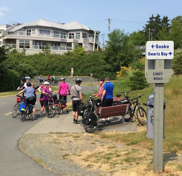 A group of eight cyclists stopped at the junction of the Galloping Goose and Lochside Regional Trails chatting with one another, with a wooden sign in the foreground with one arrow pointing to Sooke and another pointing to Swartz Bay.