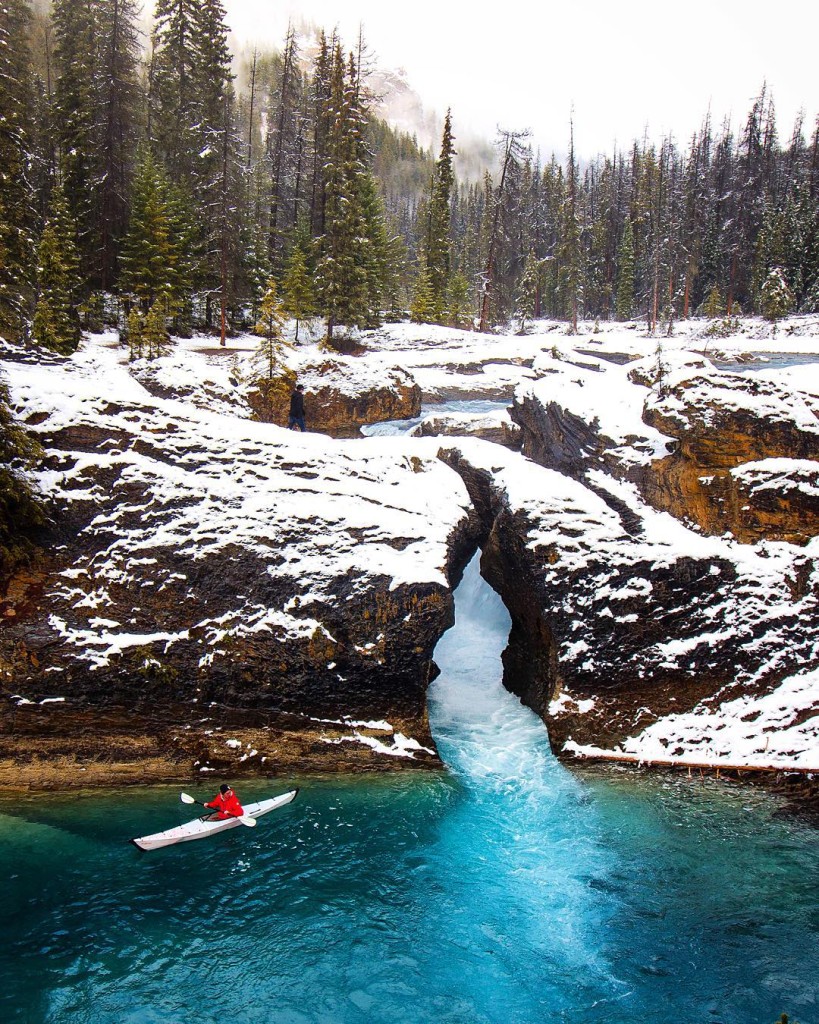 A person in a red jacket paddles a white kayak on a glacier lake by the Natural Bridge in Yoho National Park 