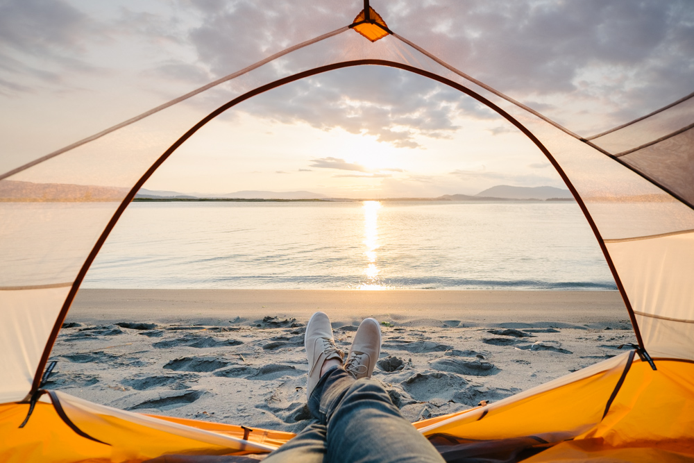 A person watches the sunset on Sidney Island on the beach. Their feet are seen at poking out the edge of their orange tent on the sand. 