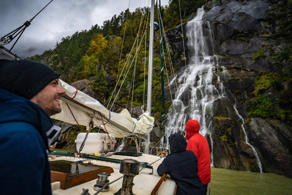 Three people on a sail boat looking up at a waterfall on an overcast day. 
