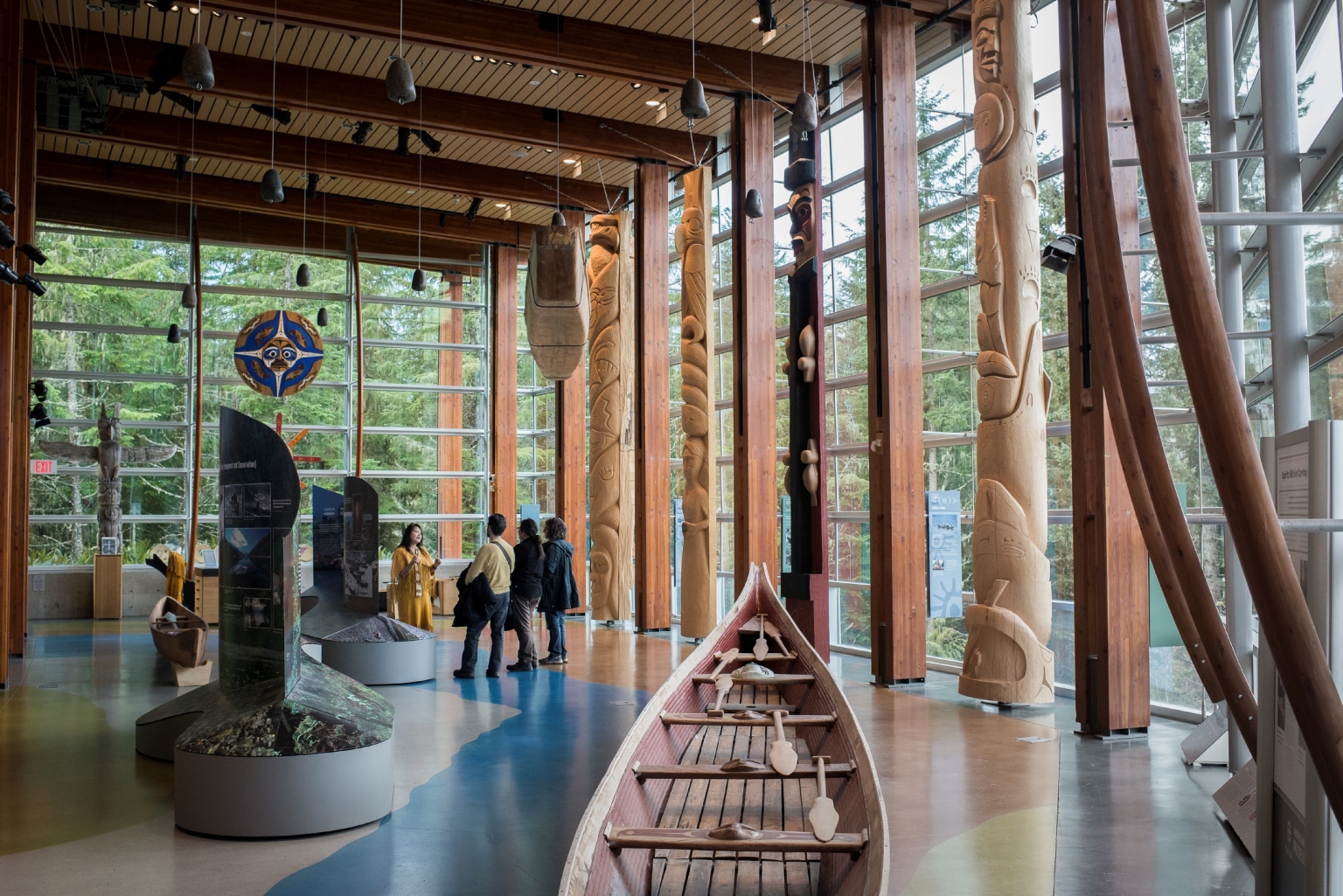 An Indigenous guide speaks to two visitors in an open gallery with cedar beams and tons of natural light. A wooden canoe sits in the foreground to the right, with totem poles and other carvings behind.