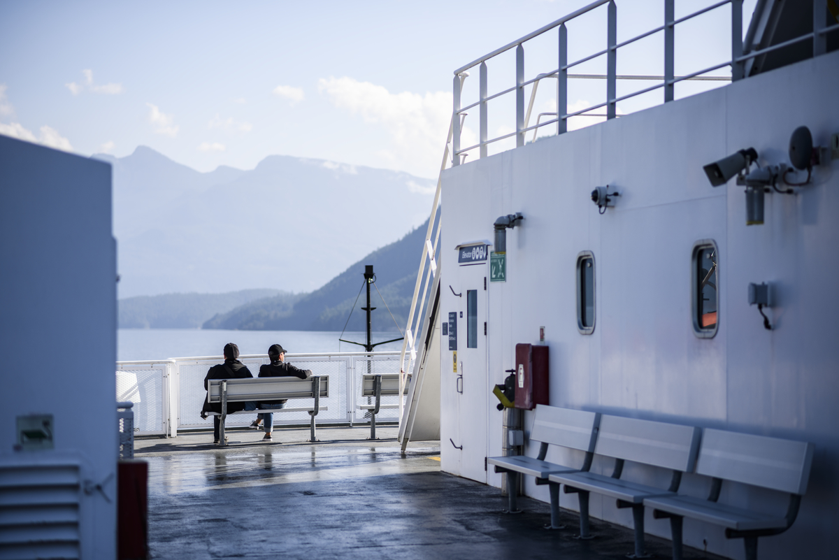 Two people sit on a ferry deck looking out at the water on a clear sky day. Mountains are seen in the distance on the other side of the ocean.