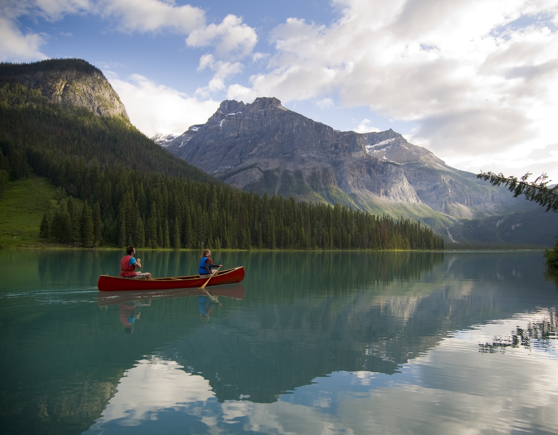 A red canoe is paddled by two people across a lake in Yoho National Park. The turquoise lake is surrounded by mountains and trees.