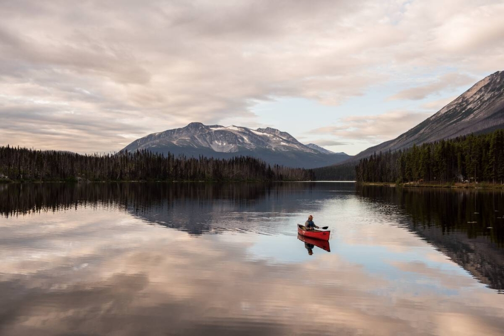 A person paddles in a red canoe on a still lake at sunset in Tweedsmuir Provincial Park