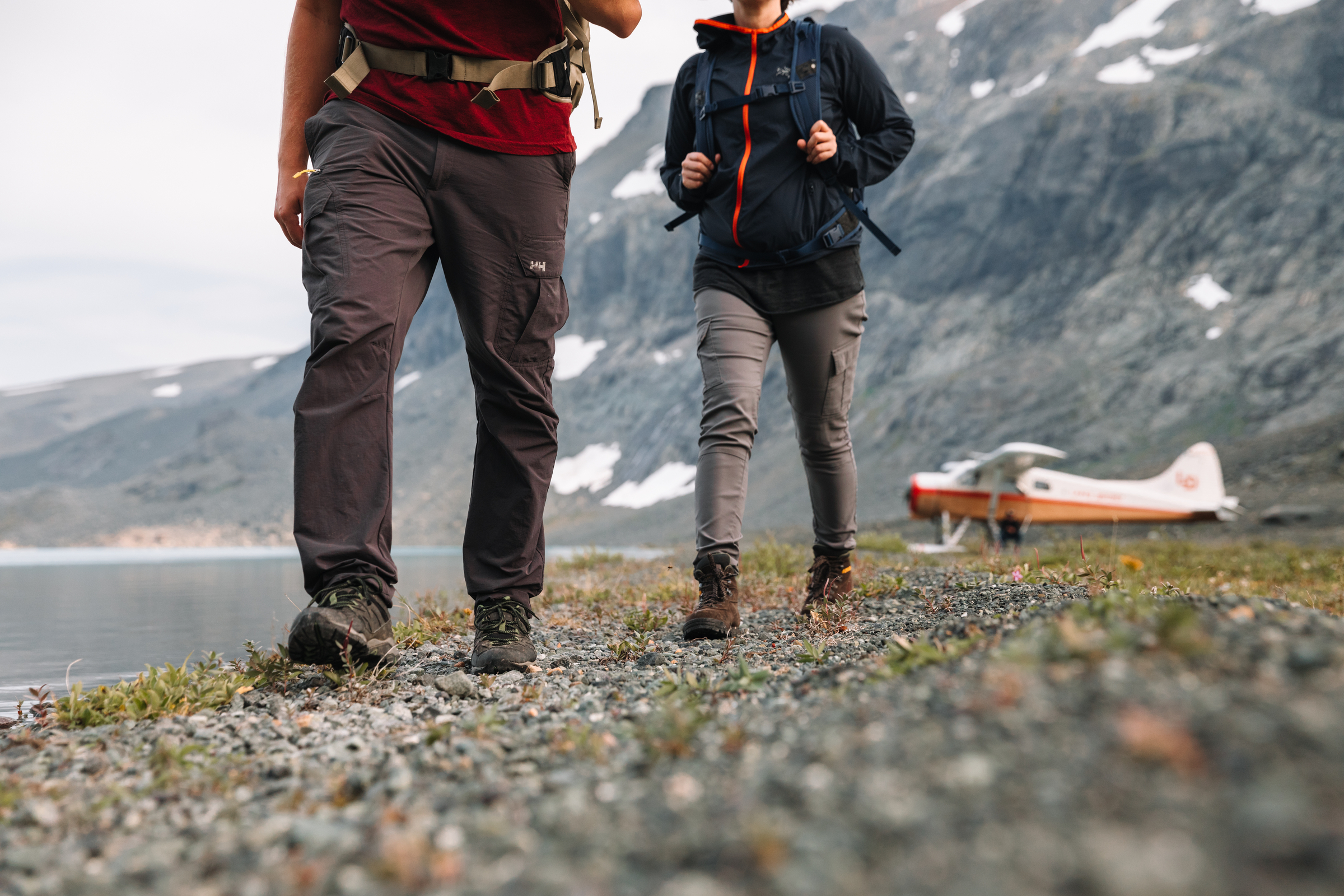 Two people walk on a lakeside trail with towering mountains and a float plane in the background.