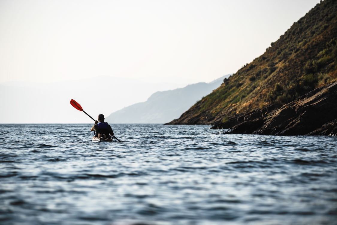 A person kayaks in calm waters off a hilly shore.
