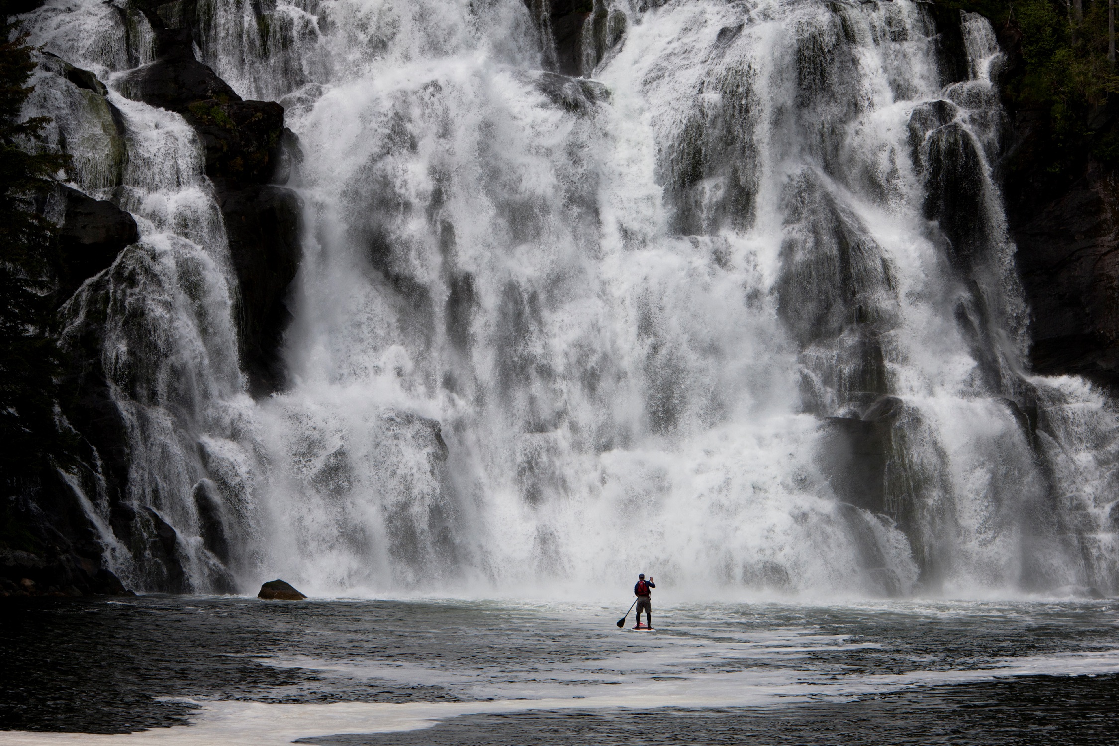 Man in the distance paddleboarding towards a cascading waterfall