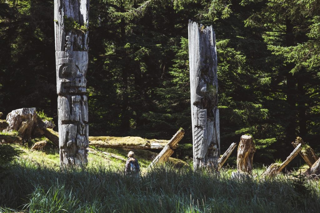 A woman standing between two old totem poles.