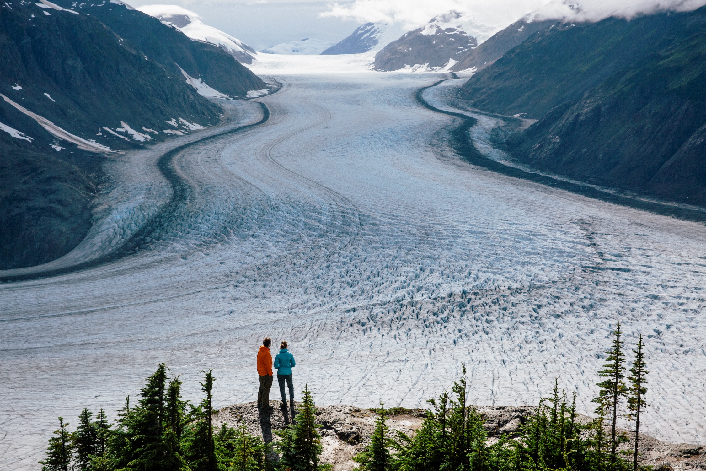 Two people look out at a frozen landscape in the Stewart-Cassiar region of northern BC. They stand on a rocky outcrop and are looking towards the mountain range.