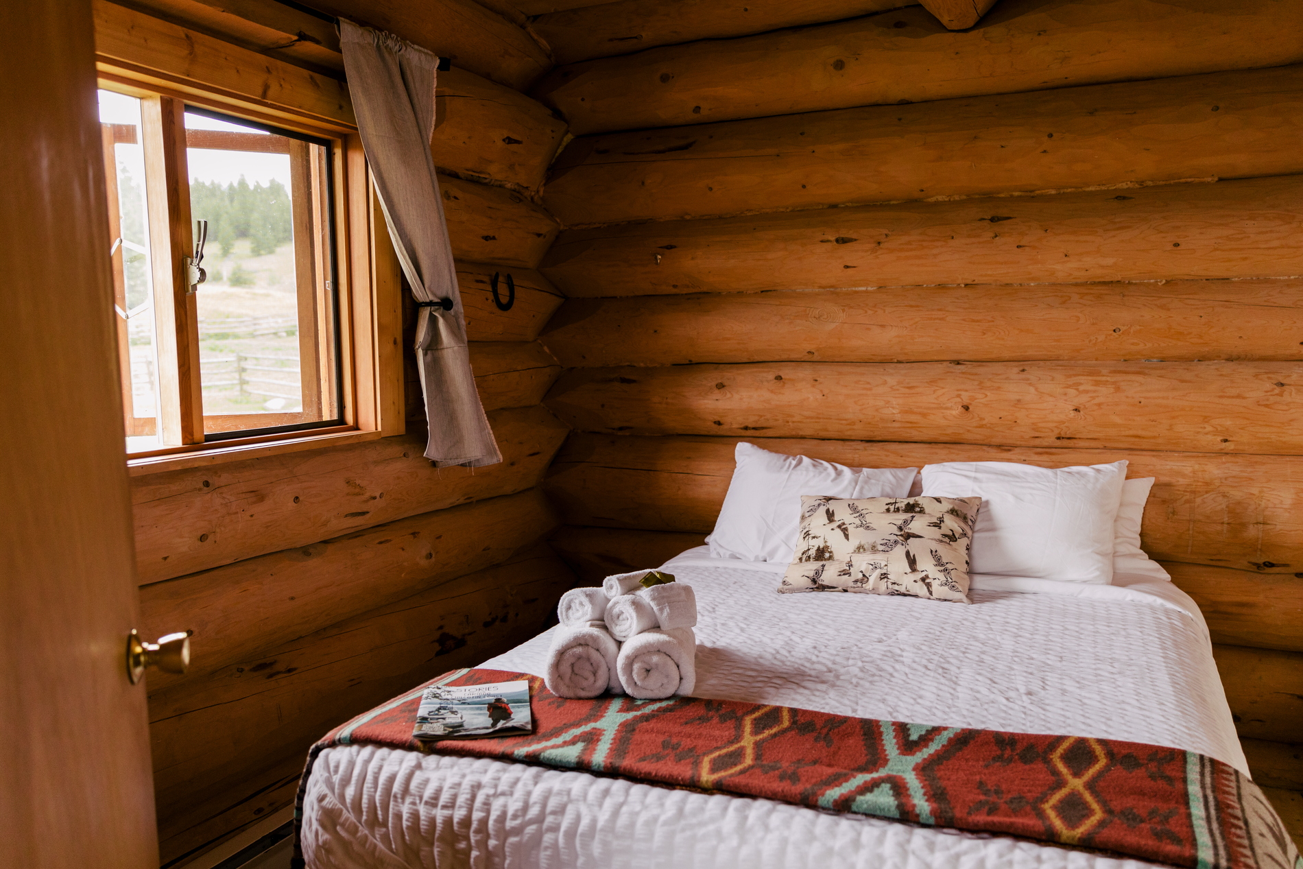 A queen size bed inside a log cabin at the Big Bar Guest Ranch. A window next to the bed is open letting in the air and light.