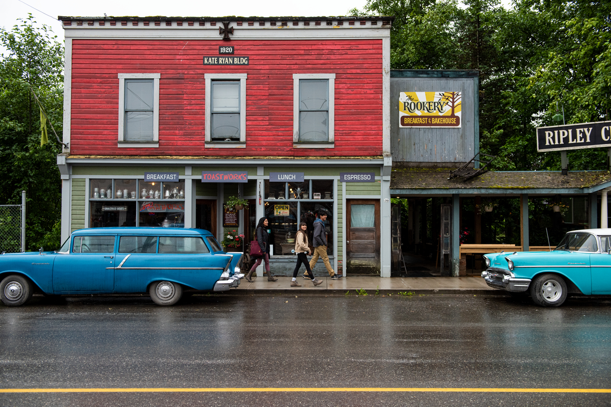 Family walking through the town of Stewart on a rainy day. Cars are parked on the side of the road in front of a diner. 
