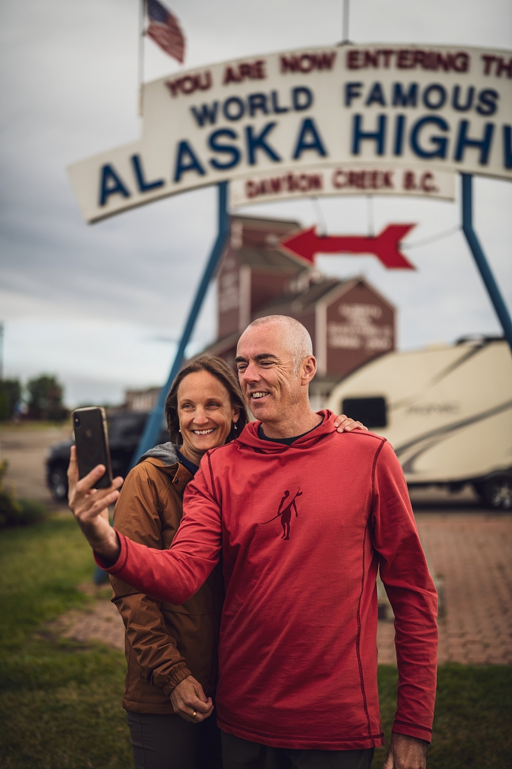 A couple takes a selfie with the Alaska Highway's Mile Zero sign in the background.
