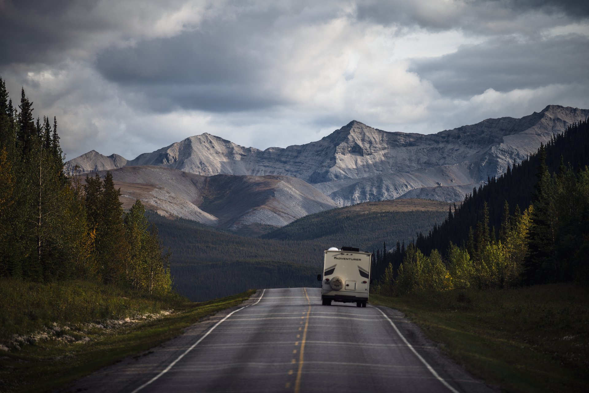 An RV drives down a quiet highway towards a mountain range in the distance. Words on the back of the RV read, "Adventures."
