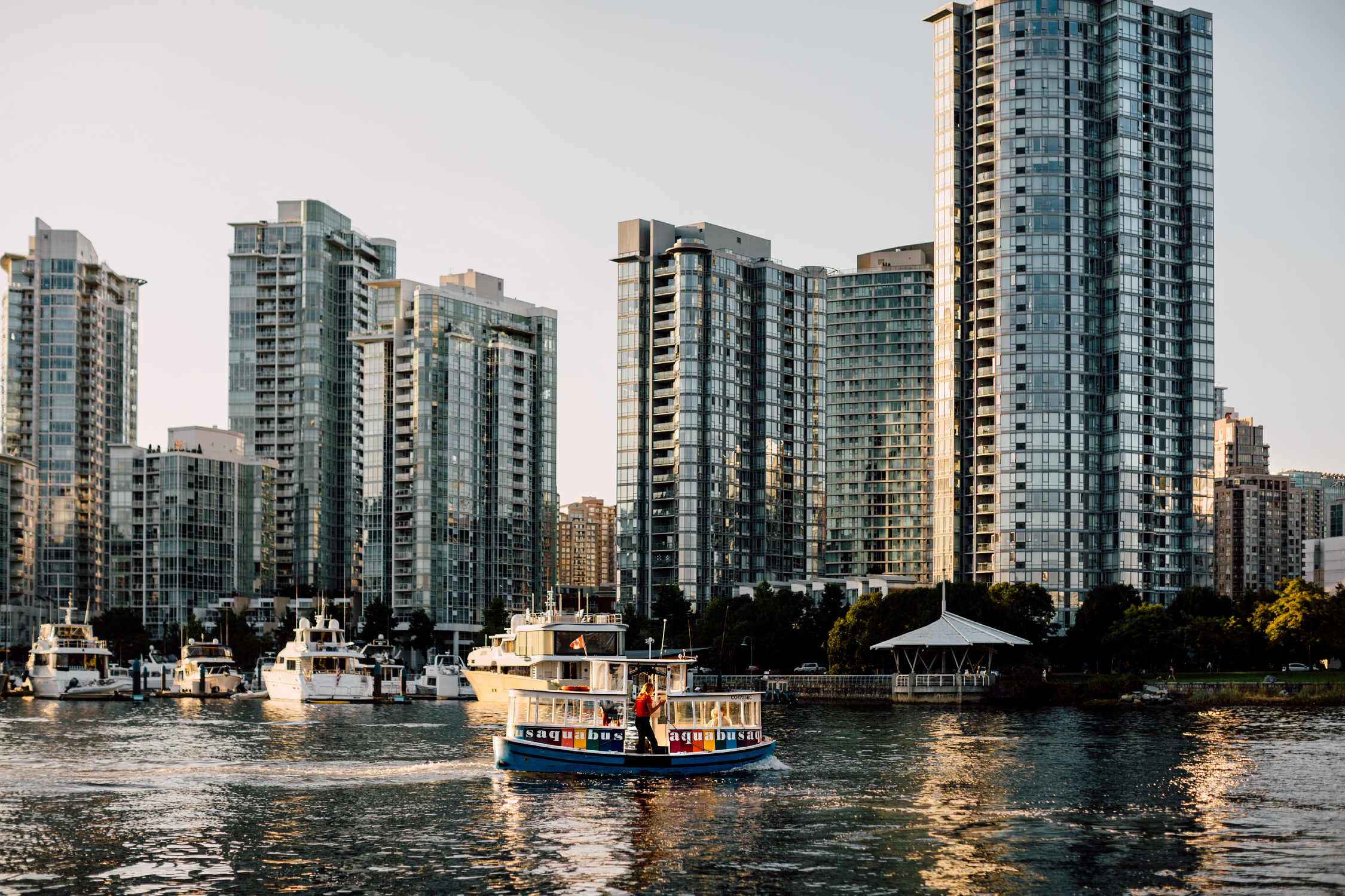 An Aquabus moves across the water from downtown Vancouver. Glass high rises and boats docked at the marina are in the background.