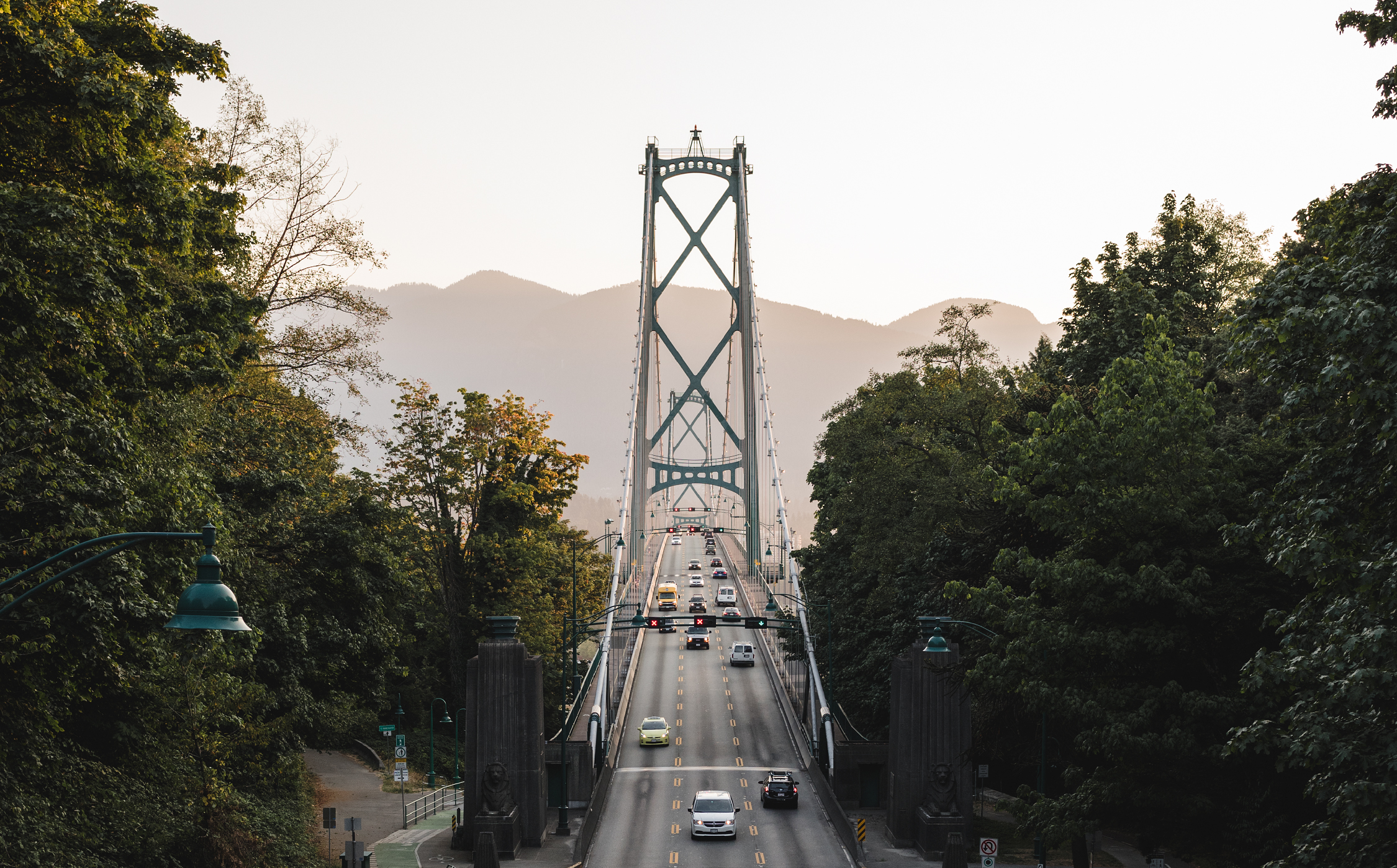 The Lion's Gate Bridge in Vancouver's Stanley Park at sunset. Cars make their way across the bridge. Large green trees surround the bridge on both sides and a glimpse of the north shore mountains are in the background.