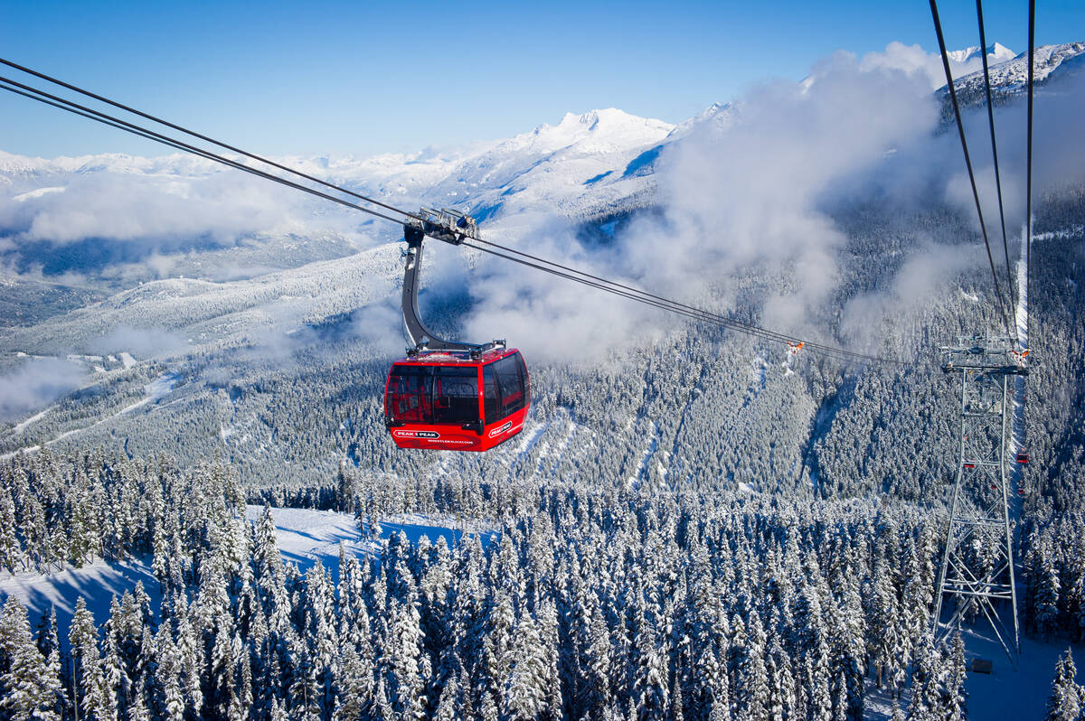 A red gondola car hangs over a snowy mountain valley with a cloudy sky and mountains behind the clouds