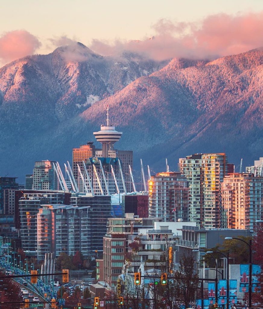 Tall buildings in downtown Vancouver stand in front of snowy mountains in the distance. The sunset is casting a pink hue onto the mountains.