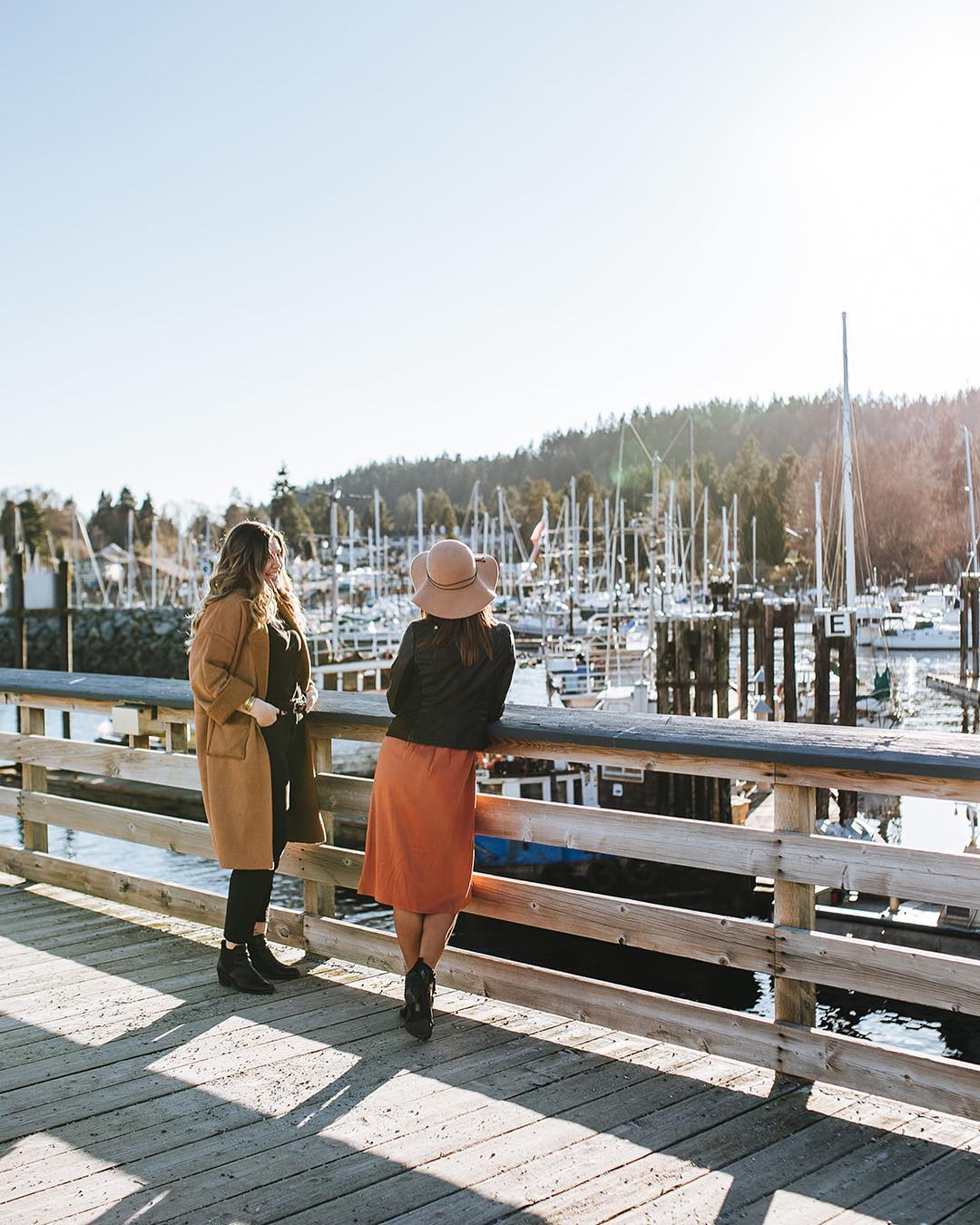 Two people stand on a pier looking out at the boats in the harbour at Gibsons Marina on a sunny day.