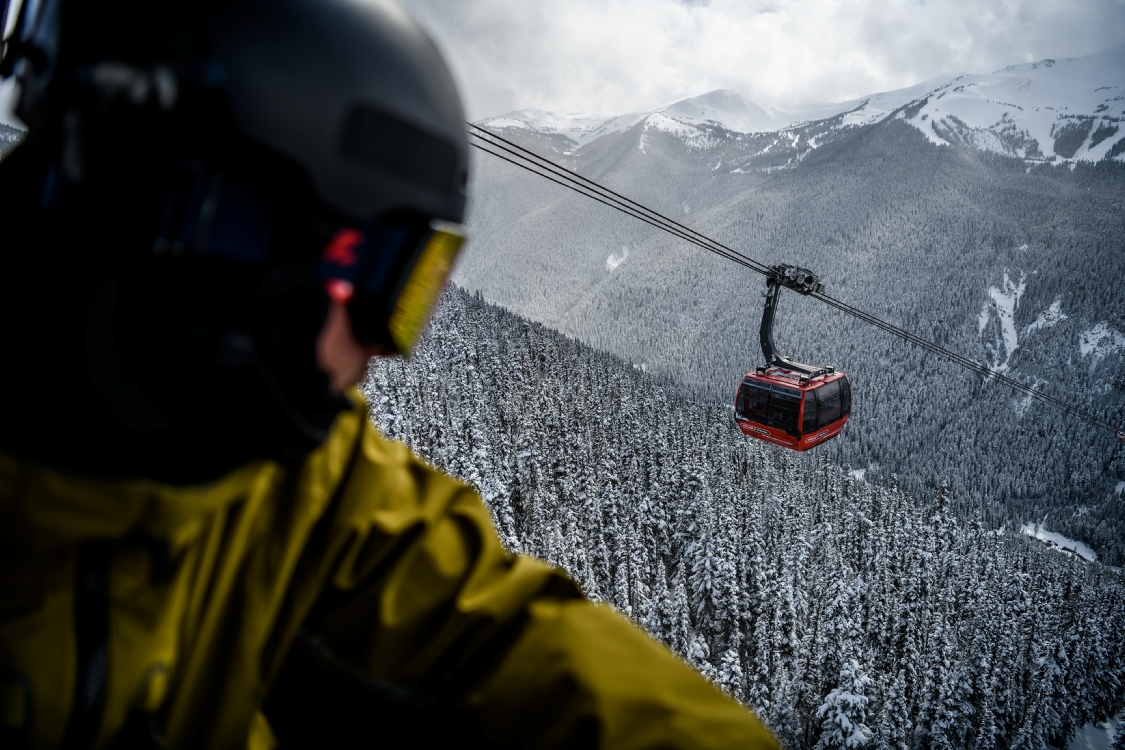A skier looks down on a passing gondola car that sits high above snowy trees and mountains.