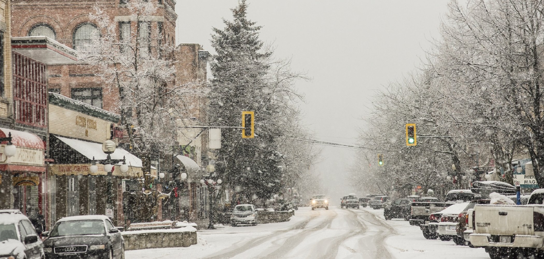 A snowy view of downtown Nelson. The snow covered street has cars on both sides. Shops line the streets.